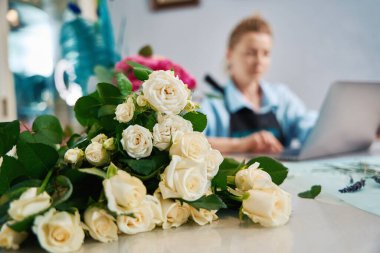 Close up of a bouquet of small roses. In the background, a female florist is working on a laptop