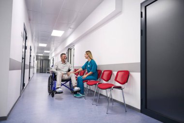 Smiling man in a wheelchair communicates with an attentive woman doctor in bright and spacious corridor of an outpatient clinic