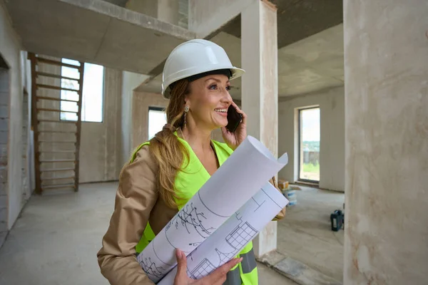 Happy Female Real Estate Agent Talking Mobile Holding Rolled Blueprints — Stockfoto