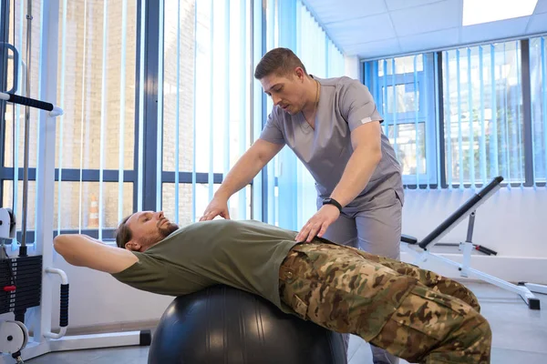 Attentive energetic doctor is engaged in physiotherapy exercises with a patient in camouflage pants on a fitball in rehabilitation room
