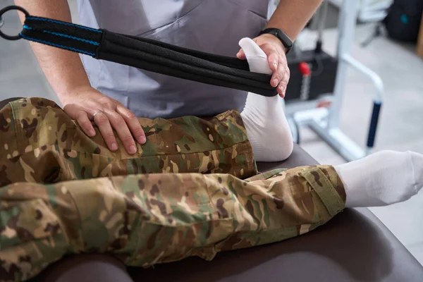 Rehabilitation specialist in a modern military hospital works with patient foot in camouflage pants and white socks on special simulator