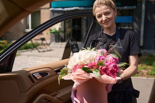 Friendly female florist gives a stylish bouquet of flowers while standing near the open car door. Composition of flowers