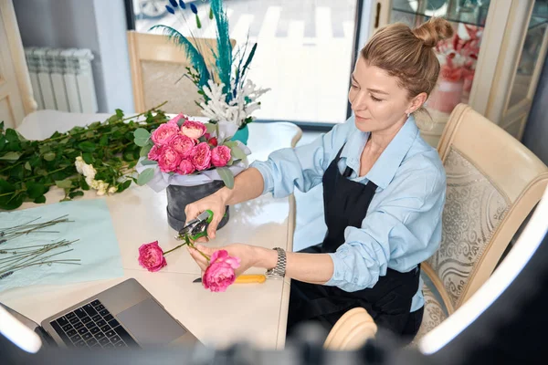 Adorable florist teaches to create stylish flower bouquets in online class. Portrait of woman sitting at table next to laptop