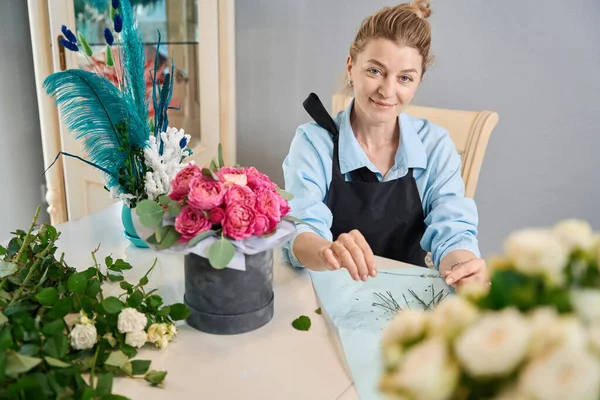 Portrait of happy florist sitting at table with trendy bouquet in box. Recording online lesson on creating compositions from flowers