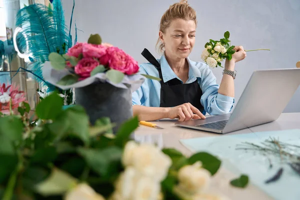 Charming woman sits in front of laptop and shows rose branch. Master class from florist on creating bouquets in box