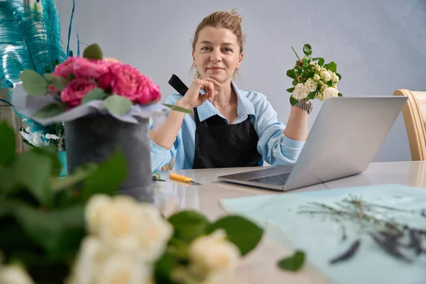 Pleasant female sits in front of laptop and demonstrates rose branch. Online lesson from florist on creating bouquets of flower