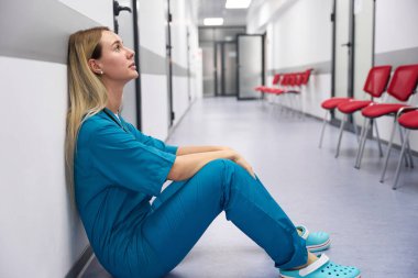 Doctor sits on the floor in the corridor leaning against the wall against the background of red chairs