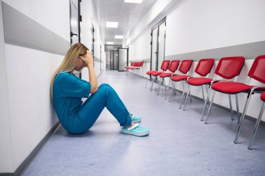 Doctor sits on the floor with his head clasped against the backdrop of a bright large corridor