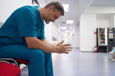 Upset unshaven doctor sitting on a red chair in a bright hospital corridor