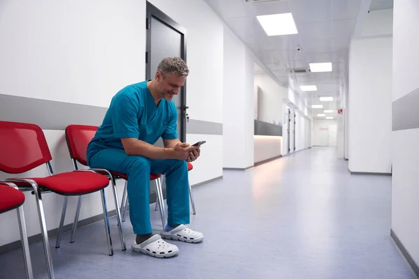 Doctor Sits Bright Corridor Looks Phone Smiling — Stockfoto