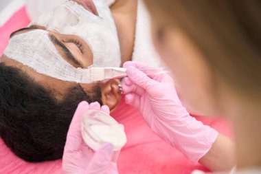 Cosmetologist in nitrile gloves applying with brush kaolin clay mask on young person face