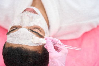 Beautician hand in nitrile glove spreading white clay paste over client face with cosmetic brush