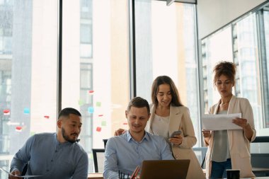 Women employees stand near sitting men and look at laptop on table. Teamwork in office with large windows