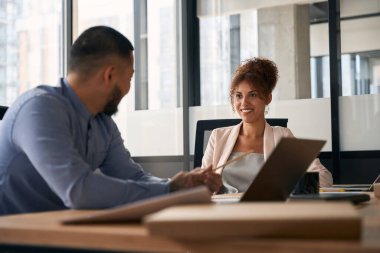 Young man and woman are talking and laughing while sitting at workplace in a spacious office. Work in large company