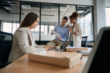 Young female sitting at laptop while co-workers are discussing documents while standing near table in office