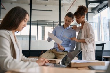 Smiling man and woman discussing working document while colleagues work on laptop