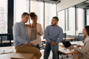 Young happy men and women who work in a modern office with large windows stand near the tables and talk
