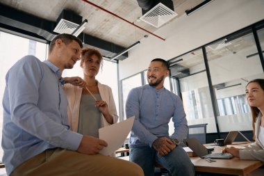 Young colleagues communicate while sitting at their desks in a spacious office with panoramic windows