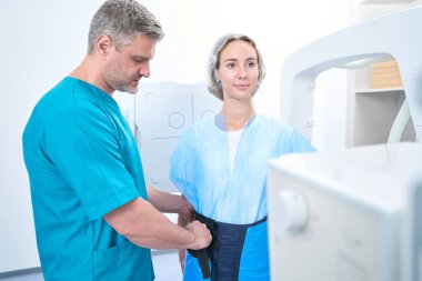 Serious radiologist at medical center putting on lead shield for x-ray procedure to smiling female patient