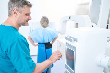 Doctor adjusts x-ray machine while female patient in lead shield waits