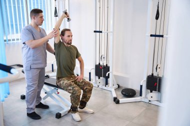 Serious physiotherapist professionally corrects the performance of therapeutic exercises with dumbbells by a soldier in the recovery period clipart
