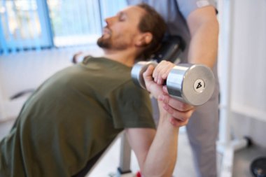 Man gently doing dumbbell exercises with the help of an experienced physiotherapist in a rehabilitation center