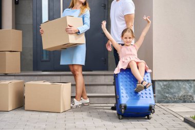 Cheerful little girl is sitting on suitcase while her parents are talking. Mom holds box with things in her hands