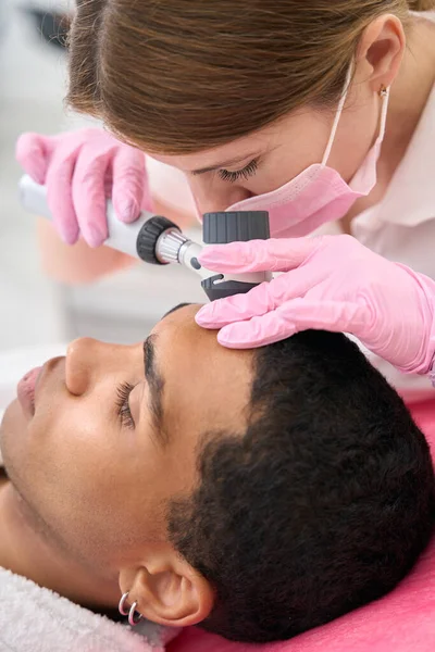 Experienced beauty clinic dermatologist examining skin on young man forehead with dermatoscope