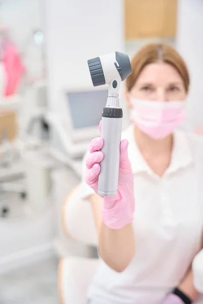 Beauty clinic female doctor in face mask and nitrile gloves holding dermatoscope before her