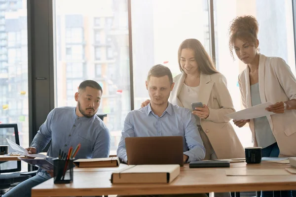 Women employees stand near sitting men and look at laptop on table. Teamwork in office with large windows