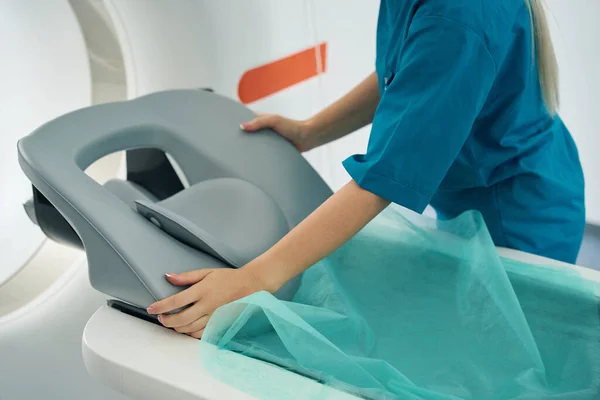 Nurse in a medical center in a special uniform covers the tomograph couch with a disposable sheet