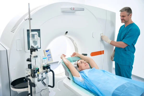Male diagnostician and female patient lying with arms outstretched up in CT scan room in medical facility