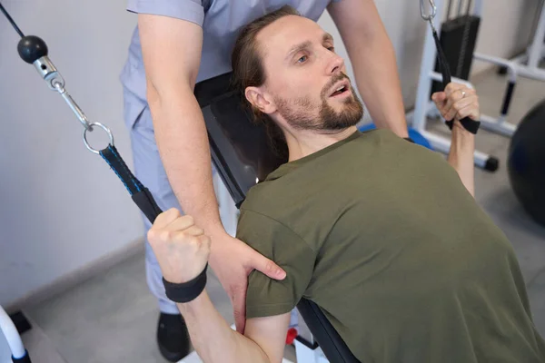 Young weakened man carefully works out on special equipment under the supervision of doctor in the office of physiotherapy exercises