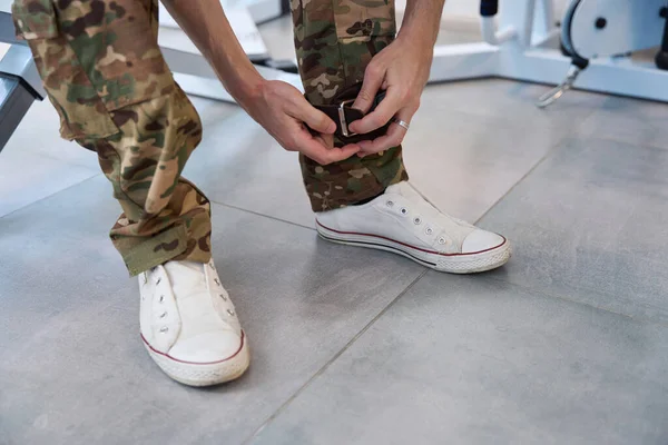 Man in camouflage clothes and training shoes puts on his ,leg a special fixing leather cuff for exercising on simulator