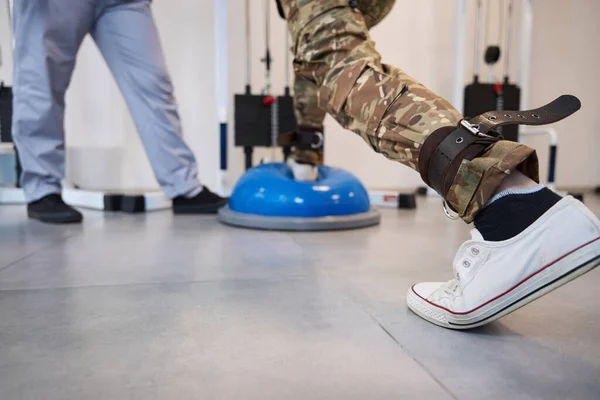 Session of restorative physiotherapy exercises in a military hospital, patient performs exercises on special equipment under the supervision of doctor