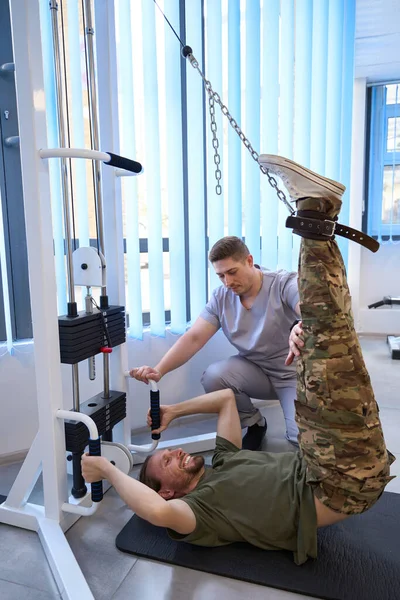 Satisfied patient undergoing spinal traction procedure after military injury on special apparatus under supervision of physiotherapist in a rehabilitation center