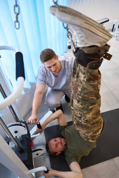 Man in camouflage clothes undergoing spinal traction procedure after military injury under the guidance of experienced instructor in rehabilitation center