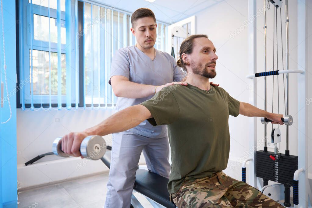 Exhausted patient diligently performs special exercises with dumbbells under the supervision of an instructor in a rehabilitation room