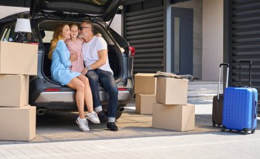 Cute daughter hugging smiling mother and father sitting in car with open trunk. Unloaded boxes and suitcases with things
