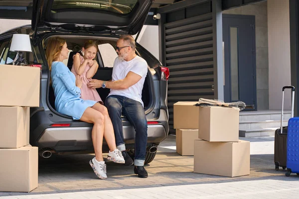 Husband and wife sit in a car with an open trunk and tickle their daughter. Move-in boxes unloaded