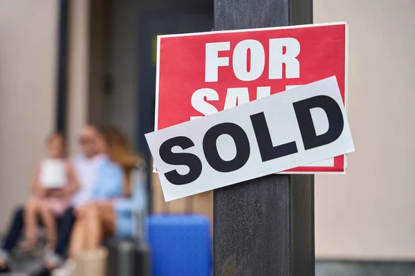 Close up sign for sale. House is sold. family sitting on the steps on the background