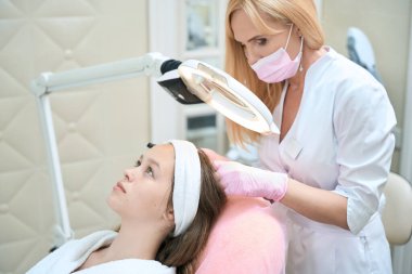 Focused beautician in salon looks through a magnifying glass lamp at the hair of young client in pink chair