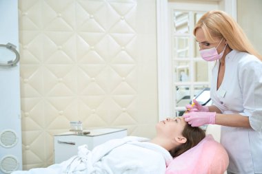 Photo of an attractive female cosmetologist in a pink mask and gloves, which makes an injection for hair growth to a girl on a pink chair