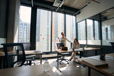 Pretty female colleagues are chatting while being in a spacious trendy office with a large window
