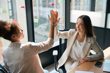 Happy female colleagues give each other high five while being in coworking space with big windows and beautiful view