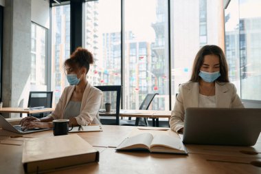 Pretty young women in protective masks work in the office, sitting at laptops against the background of a panoramic window
