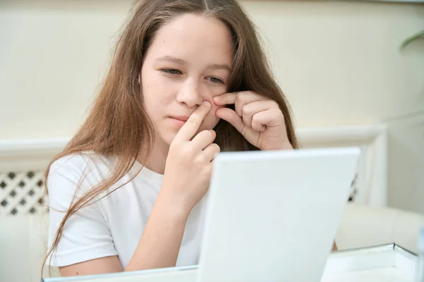 Young girl looks in the mirror on the table and squeezes pimples under her eye with both hands