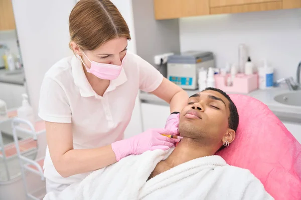 Focused dermatologist in nitrile gloves injecting dermal filler into wrinkle on young man neck