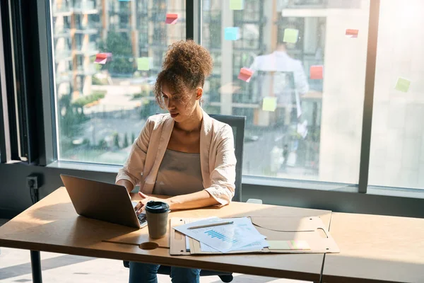 African american woman works at laptop in office against background of majestic window with beautiful view