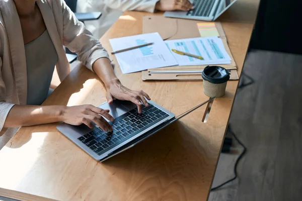 Cropped photo of woman typing on computer in coworking space. There is cup of coffee and documents on table nearby